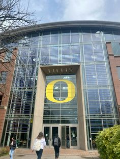 two people walking in front of a large building with the letter o on it's side