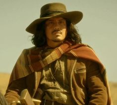 a man with long hair wearing a hat and scarf on top of a horse in the desert