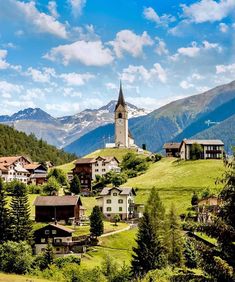 a small village on the side of a mountain with a church in the middle and snow capped mountains behind it