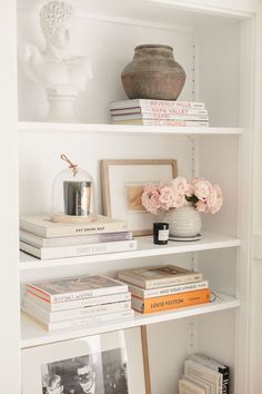 a white book shelf filled with books and vase on top of each shelf in front of a busturine