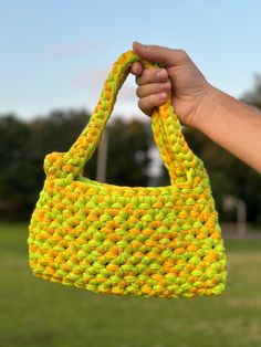 a hand holding a yellow and green bag in front of a grassy field with trees