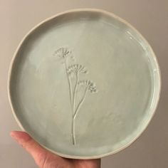 a hand holding a white plate with three flowers etched on the front and sides,