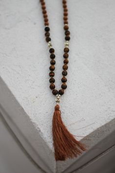 this 108 bead mala is designed by here & there creative and handmade in northern india with sandalwood and lava stone beads. the beads originate from the spiritual lands of northern india and have been blessed by the holy ganges river.  the tiger's eye beads vary in color from golden yellow to deep red. tiger's eye is a protection agent known as the "all-seeing all-knowing eye." with this mala, you'll have an added sense of security and comfort. here & there styling tip: if the tassel strands be Spiritual Brown Mala For Healing, Brown Beaded Holistic Mala, Hand-strung Brown Mala For Healing, Brown Bohemian Mala With 108 Beads, Bohemian Brown Mala With 108 Beads, Adjustable Brown Mala For Healing, Holistic Brown Beaded Mala, Spiritual Brown Mala For Meditation, Brown Spiritual Mala For Meditation