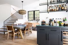 a dining room table and chairs with bottles on the shelves above it, in front of a staircase