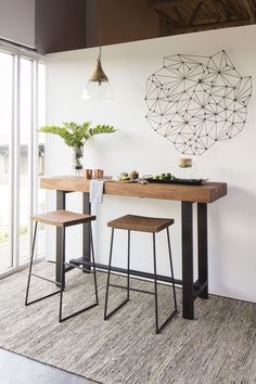 two stools and a table in front of a large window with an art piece on the wall