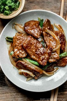 a white plate topped with meat and onions next to chopsticks on a wooden table