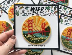 a hand holding a card with embroidered patches on it and the san francisco bridge in the background