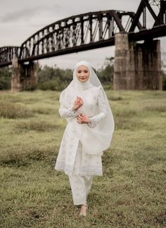 a woman standing in the grass wearing a white dress and veil with an old bridge in the background