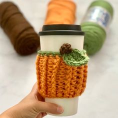 a hand holding a coffee cup sleeve in front of three yarn spools