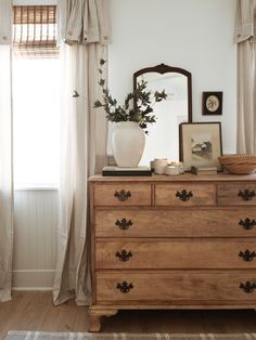 a wooden dresser sitting next to a window with drapes on it's windowsill