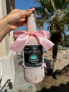 a person holding up a pink bottle with a bow on it's neck and palm trees in the background