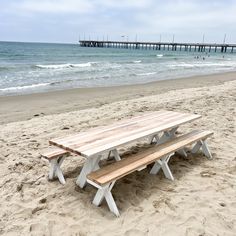 a wooden picnic table sitting on top of a sandy beach next to the ocean with a pier in the background