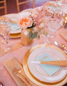 the table is set with gold and white plates, silverware, and pink flowers