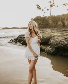 a beautiful blonde woman standing on top of a beach next to the ocean wearing a white dress