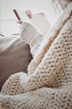 a woman reading a book while sitting on a couch with her arm wrapped in a blanket