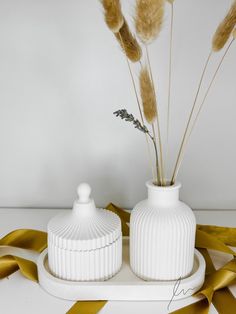 two white vases sitting on top of a table with gold ribbons around them and some dried plants in the middle