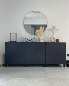 a large round mirror on the wall above a black sideboard with vases and plants