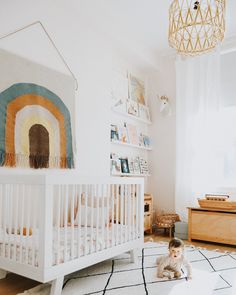 a baby sitting on the floor in front of a crib with a rainbow wall hanging above it