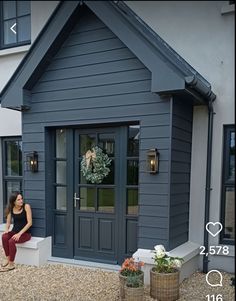 a woman sitting on a bench in front of a gray house with wreaths and potted plants