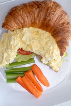 a croissant sandwich with eggs and carrots on a white plate next to celery