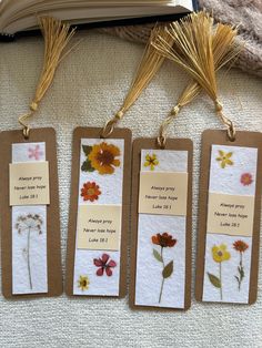 three bookmarks with flowers on them sitting next to an open book and some grass