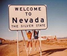 two people standing on a welcome to nevada sign in front of a dirt road and desert area