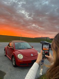 a woman taking a photo of a red volkswagen beetle parked in a parking lot at sunset