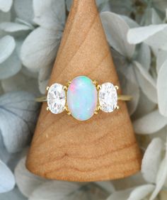 an opal and diamond three stone ring sitting on top of a wooden stand surrounded by flowers