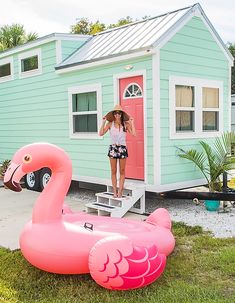 a woman standing next to a pink flamingo in front of a tiny blue house