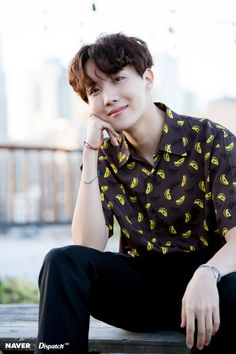 a young man sitting on top of a wooden bench wearing a banana shirt and black pants