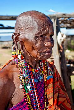 an old woman with beads and necklaces on her neck standing in front of a wooden structure