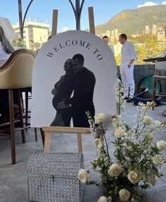 a couple kissing in front of a welcome sign with flowers on the ground next to it