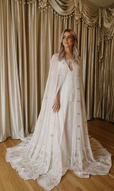 a woman standing in front of curtains wearing a white gown and cape with lace trims