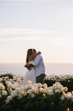 a bride and groom kissing in the middle of white flowers by the ocean at sunset