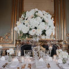 a table set with white flowers and candles in front of a fire place decorated with gold trimmings