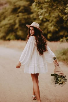 a woman in white dress and hat walking down the road with her hand on her hip