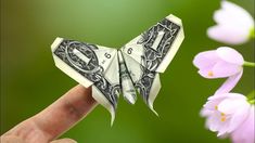 a dollar bill origami butterfly sitting on someone's finger with pink flowers in the background