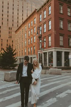 a man and woman walking across a street in front of tall buildings on a sunny day