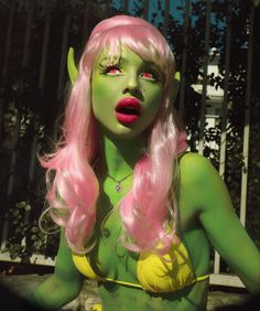 a woman with pink hair and green makeup is posing for a photo in front of a fence