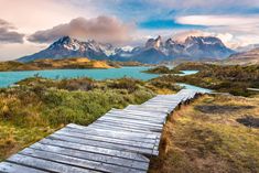 a wooden walkway leading to a lake with mountains in the background and clouds above it