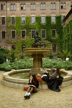 two women sitting on the ground in front of a fountain with ivy covered buildings behind them