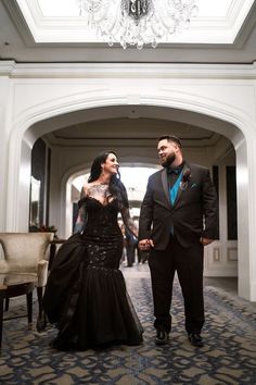 a man and woman dressed in formal wear walking down a hall way with chandelier hanging from the ceiling