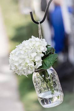 white flowers are in a glass jar hanging from a black cord on a hook outside