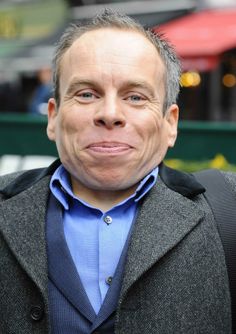 a man with grey hair wearing a blue shirt and gray jacket looking at the camera