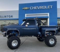 a large blue truck parked in front of a chevrolet dealership with big tires on it's flatbed