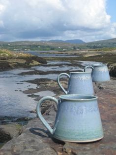 three blue mugs sitting on top of a rock next to a body of water