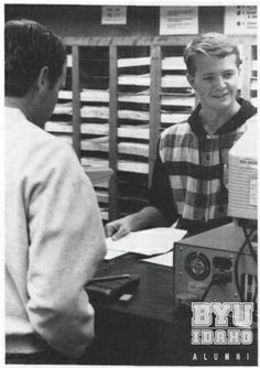 a young man sitting at a desk in front of a computer monitor and another person standing next to him