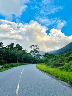 an empty road with a rainbow in the sky and trees on both sides, surrounded by greenery