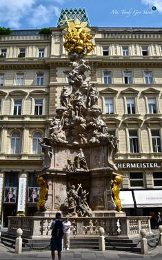 two people standing in front of a building with statues on the outside and gold decorations on the top