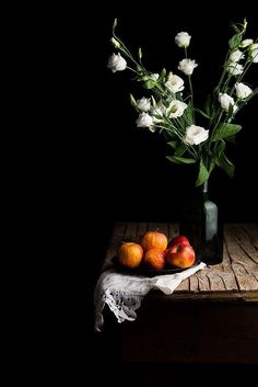 a vase filled with white flowers and fruit on top of a wooden table in front of a black background